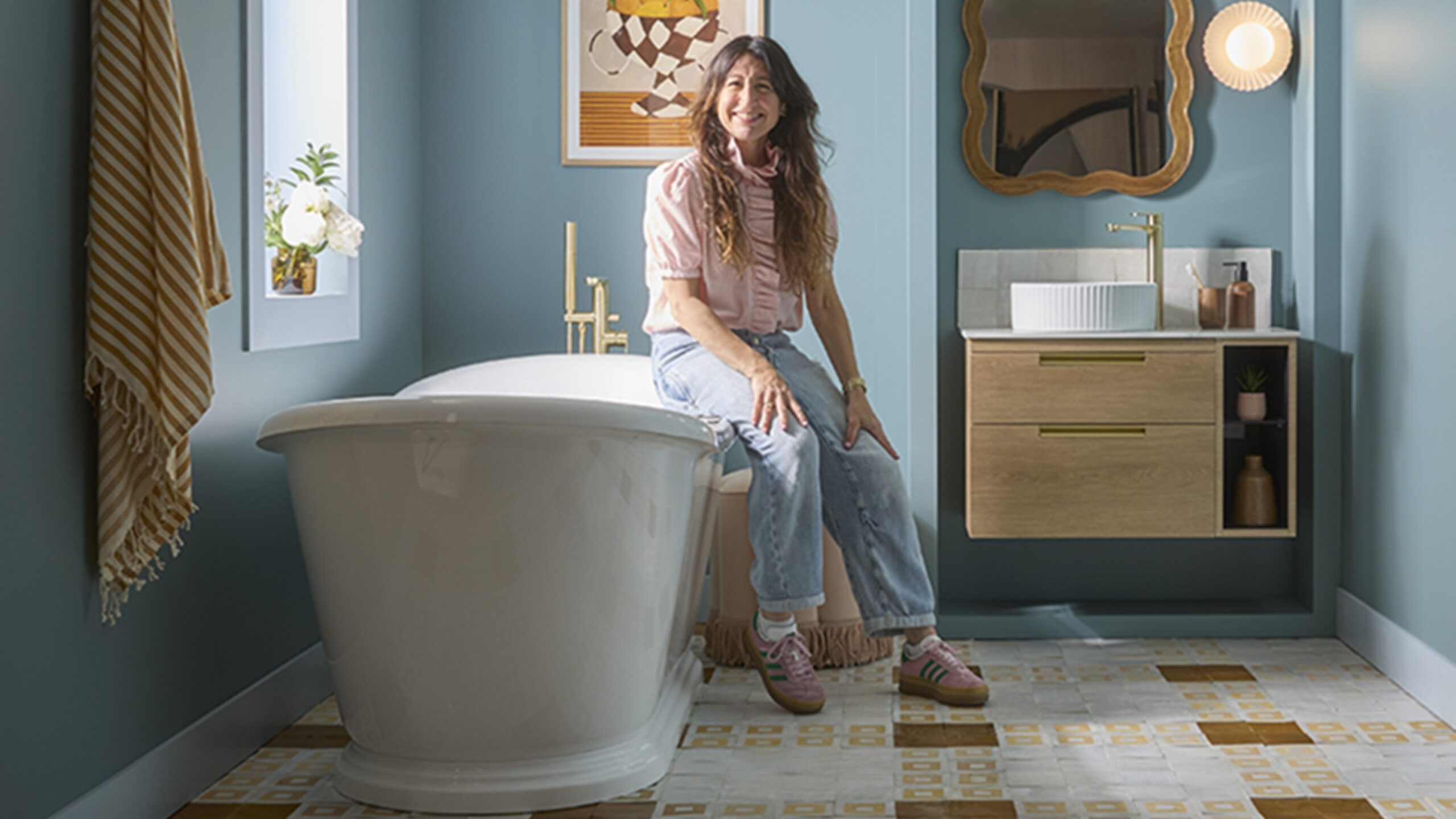 A woman with long, dark hair is seated on the edge of a white, freestanding bathtub in a bathroom. She is wearing a light-coloured, long-sleeved top and blue jeans. To her right, there is a wooden wall-mounted vanity unit with a white circular sink and a gold-coloured faucet.