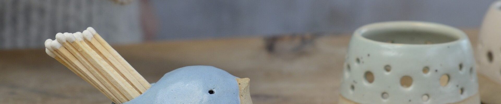 A ceramic bird-shaped match striker pot in a cornflower blue colour with a smiling face, holding several wooden matches, sits on a wooden surface next to another lighter-coloured, textured ceramic pot