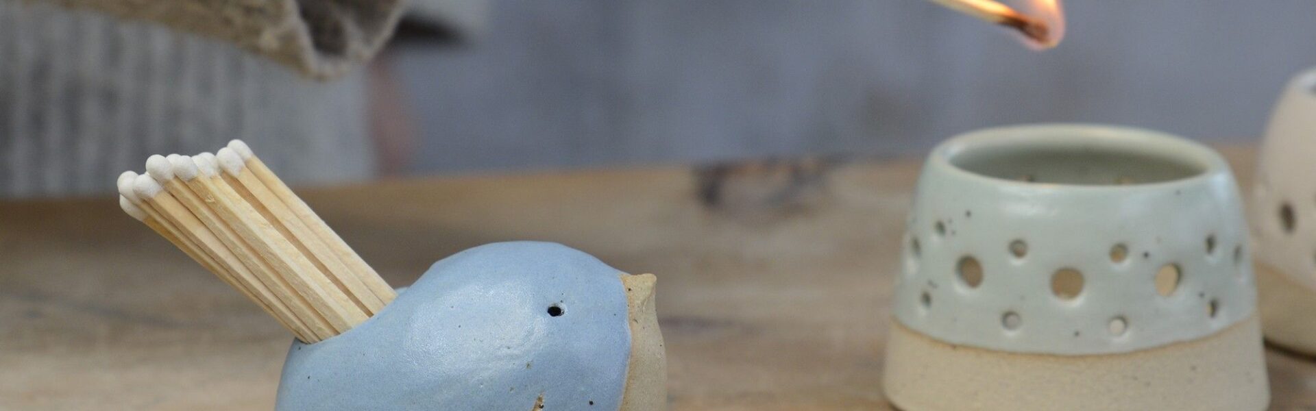 A ceramic bird-shaped match striker pot in a cornflower blue colour with a smiling face, holding several wooden matches, sits on a wooden surface next to another lighter-coloured, textured ceramic pot