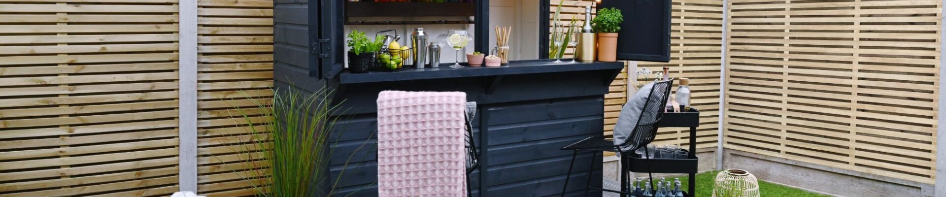 A garden bar made from a dark-painted wooden shed with a pent roof. The front opens up to reveal a bar area with shelves stocked with bottles, glasses, and bar tools. String lights are hung inside.