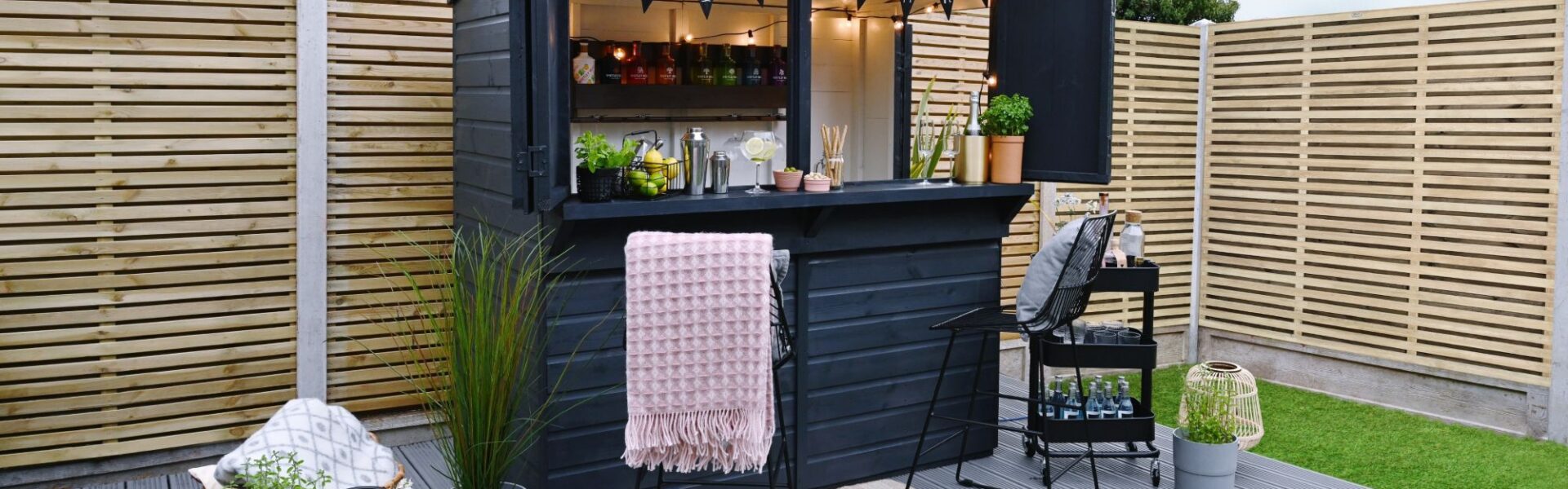 A garden bar made from a dark-painted wooden shed with a pent roof. The front opens up to reveal a bar area with shelves stocked with bottles, glasses, and bar tools. String lights are hung inside.
