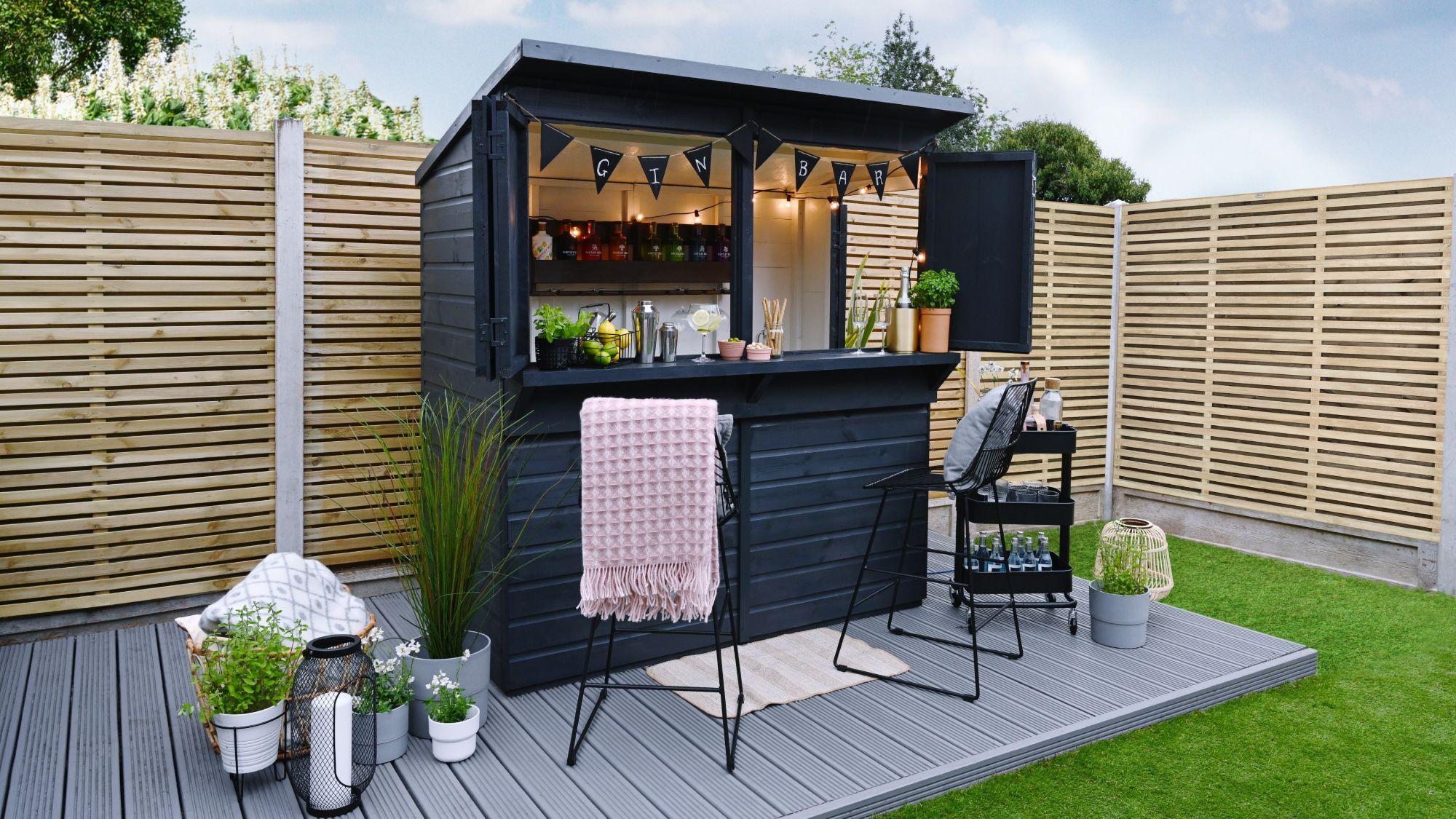 A garden bar made from a dark-painted wooden shed with a pent roof. The front opens up to reveal a bar area with shelves stocked with bottles, glasses, and bar tools. String lights are hung inside.
