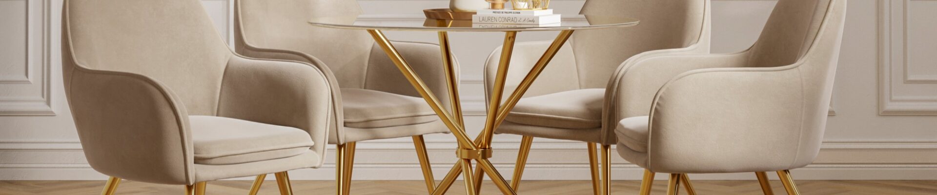 A round glass dining table with a gold base is surrounded by four beige velvet dining chairs with gold legs. The table is adorned with a white vase holding white flowers and a stack of books with some decorative items on top. The room features white panelled walls and a light-coloured wooden floor.