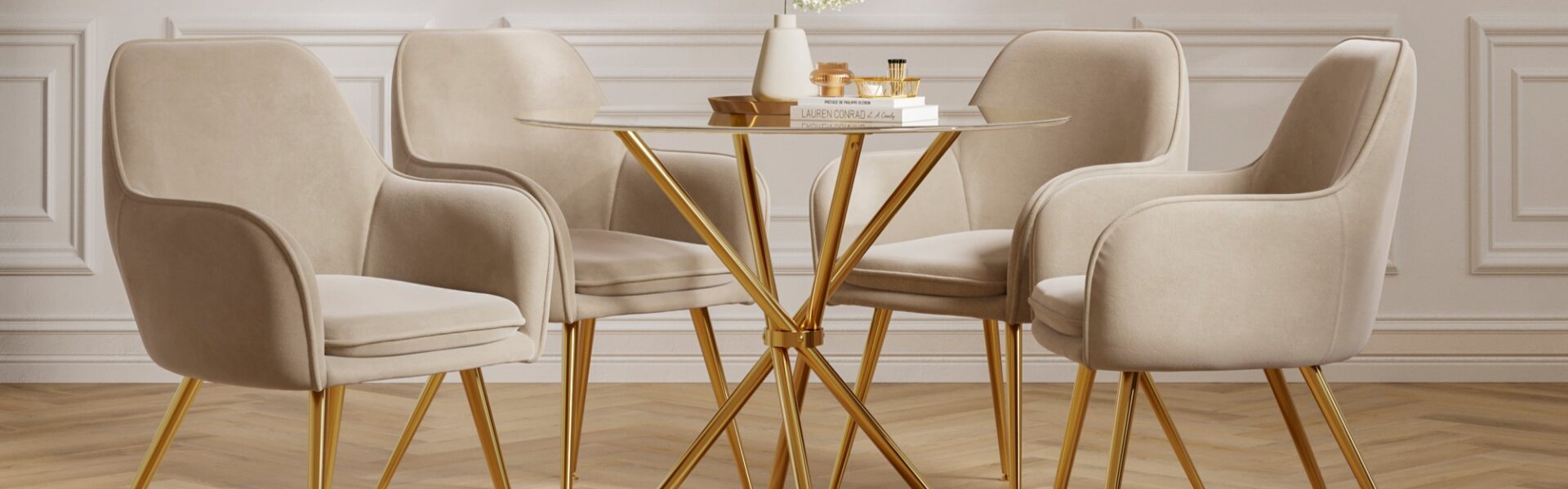 A round glass dining table with a gold base is surrounded by four beige velvet dining chairs with gold legs. The table is adorned with a white vase holding white flowers and a stack of books with some decorative items on top. The room features white panelled walls and a light-coloured wooden floor.