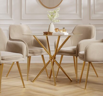 A round glass dining table with a gold base is surrounded by four beige velvet dining chairs with gold legs. The table is adorned with a white vase holding white flowers and a stack of books with some decorative items on top. The room features white panelled walls and a light-coloured wooden floor.