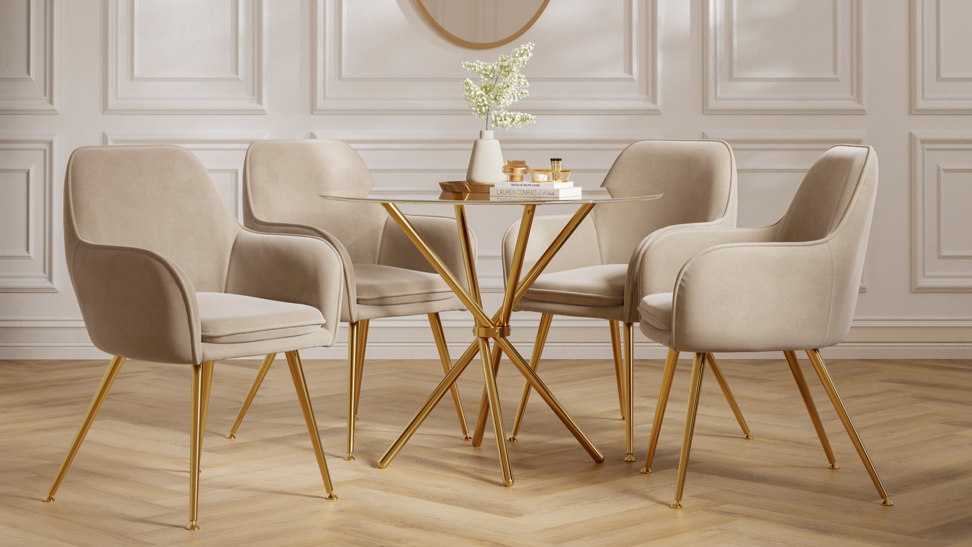 A round glass dining table with a gold base is surrounded by four beige velvet dining chairs with gold legs. The table is adorned with a white vase holding white flowers and a stack of books with some decorative items on top. The room features white panelled walls and a light-coloured wooden floor.