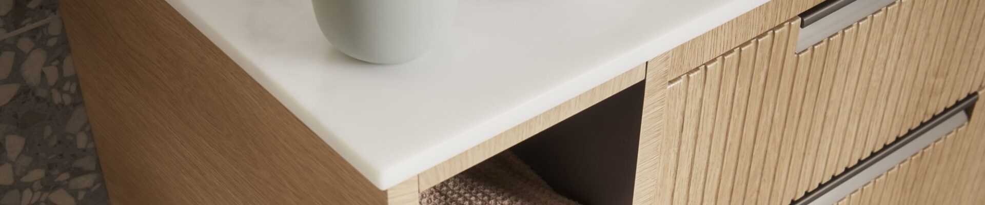 A close-up shot of a bathroom vanity unit. The unit has a light wood finish with a subtle fluted or ridged texture on the drawer fronts. It features a clean white countertop.