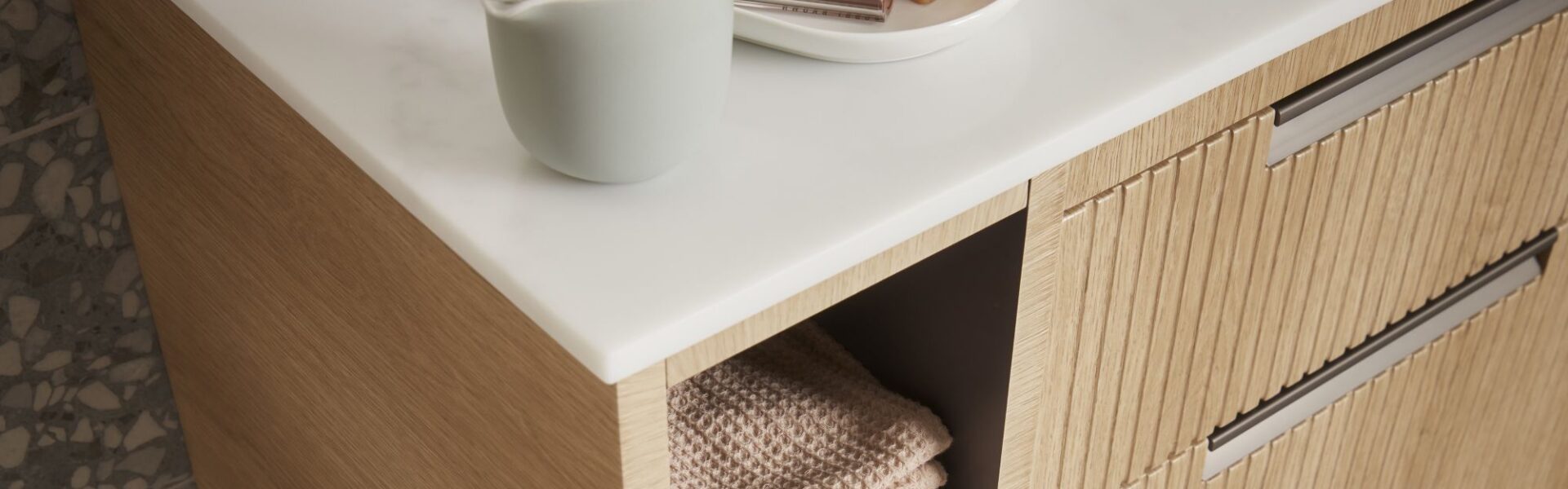 A close-up shot of a bathroom vanity unit. The unit has a light wood finish with a subtle fluted or ridged texture on the drawer fronts. It features a clean white countertop.