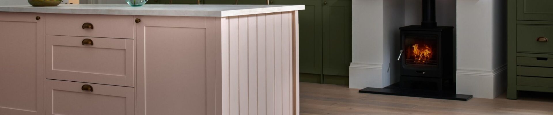 A beautifully styled kitchen features a large pink kitchen island with a white countertop and gold hardware, taking centre stage in the foreground. The floor throughout the kitchen is made of light-coloured engineered wood