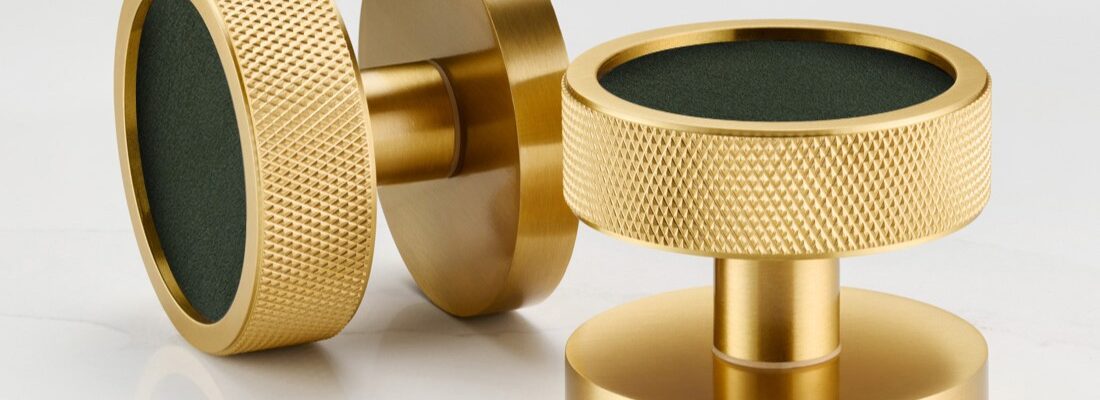 A close-up shot features a modern, round door knob in a striking design. The knob has a brass or gold-toned metallic outer ring, giving it a sleek and sophisticated look. The centre of the door knob is inlaid with what appears to be white terrazzo, speckled with various shapes and sizes of flecks in shades of black, grey, terracotta, and brown.