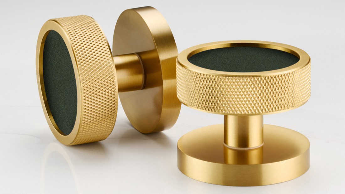 A close-up shot features a modern, round door knob in a striking design. The knob has a brass or gold-toned metallic outer ring, giving it a sleek and sophisticated look. The centre of the door knob is inlaid with what appears to be white terrazzo, speckled with various shapes and sizes of flecks in shades of black, grey, terracotta, and brown.