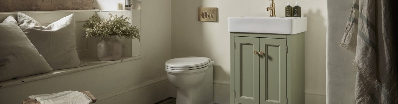 A warm and inviting bathroom features a Roper Rhodes Hampton 575 Freestanding Vanity Unit in Matt Sage Green with a white basin and gold-toned tap, positioned next to a white toilet. The vanity has two doors with round, light-coloured handles.