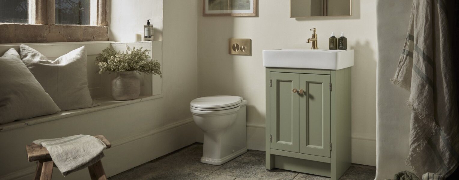 A warm and inviting bathroom features a Roper Rhodes Hampton 575 Freestanding Vanity Unit in Matt Sage Green with a white basin and gold-toned tap, positioned next to a white toilet. The vanity has two doors with round, light-coloured handles.