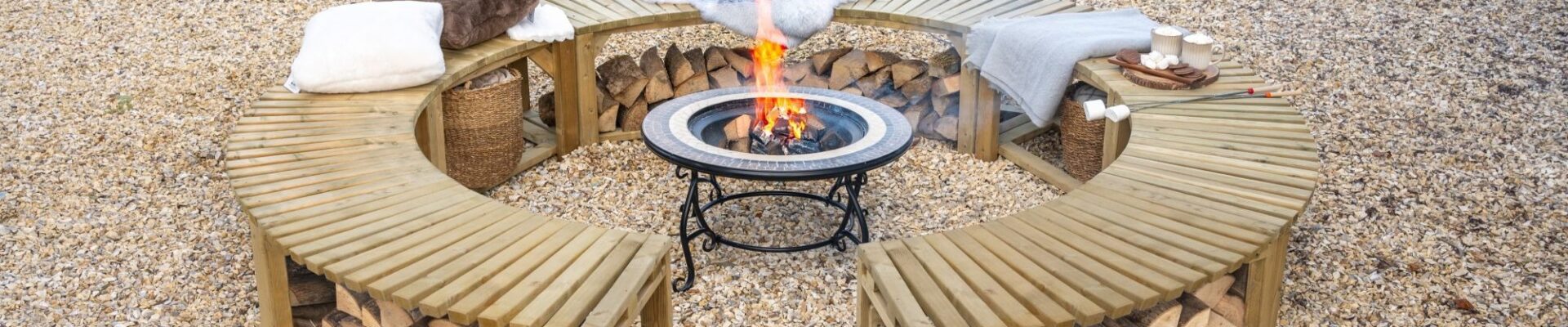 A low-angle shot shows a curved, wooden firepit bench, split into two semicircles, arranged around a central firepit. The benches are made of slatted wood and feature an under-bench shelf, which is filled with neatly stacked firewood. The benches are topped with soft cushions and blankets, suggesting a cosy and comfortable atmosphere.