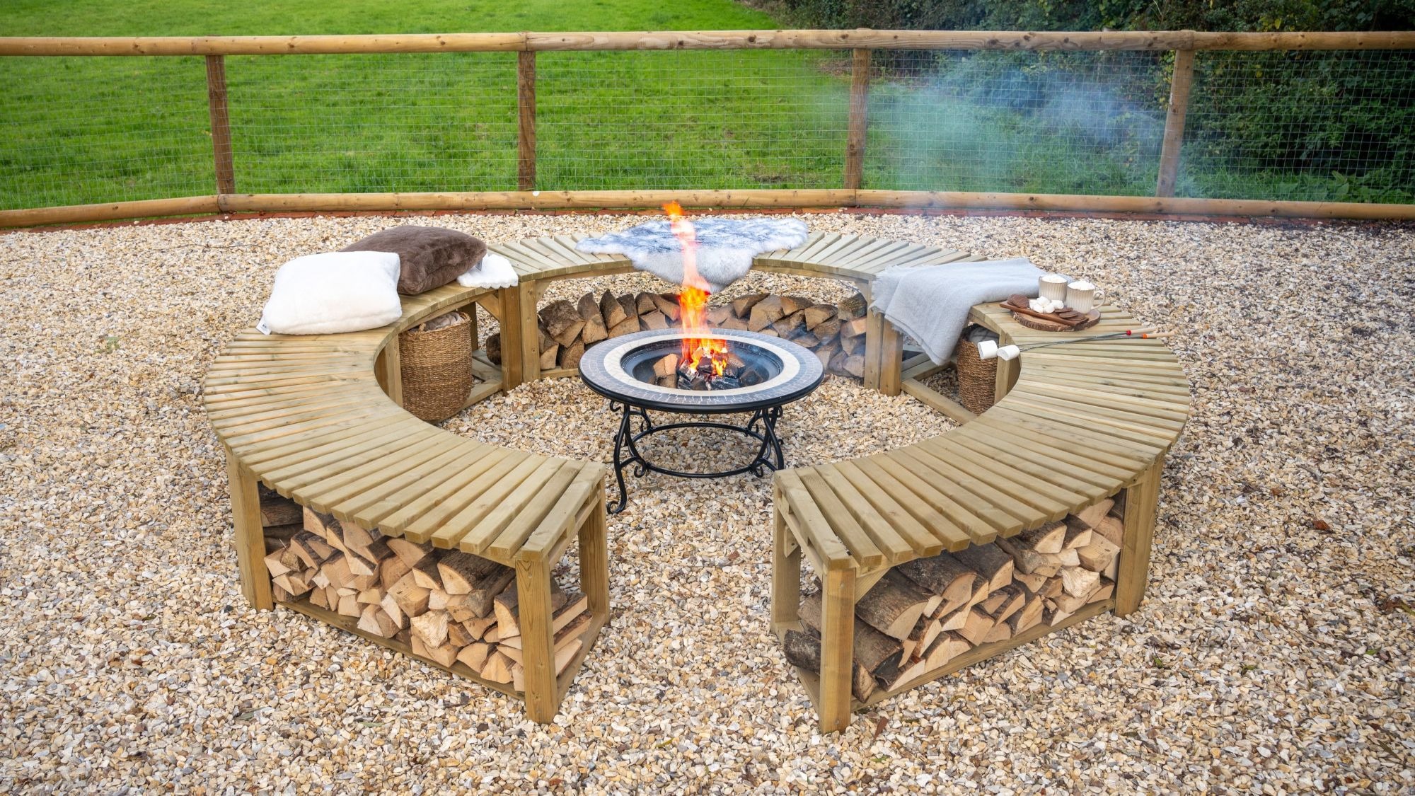 A low-angle shot shows a curved, wooden firepit bench, split into two semicircles, arranged around a central firepit. The benches are made of slatted wood and feature an under-bench shelf, which is filled with neatly stacked firewood. The benches are topped with soft cushions and blankets, suggesting a cosy and comfortable atmosphere.