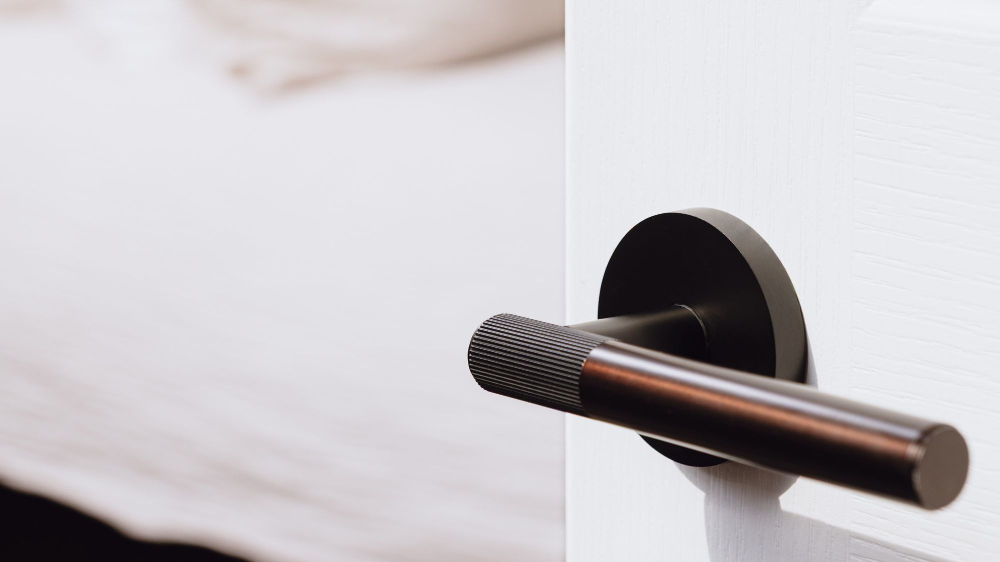 A close-up shot of a modern brass linear lever handle in a bronze finish, installed on a white door. The handle has a sleek, cylindrical shape with fine ridging near the base, and it's mounted on a circular backplate.