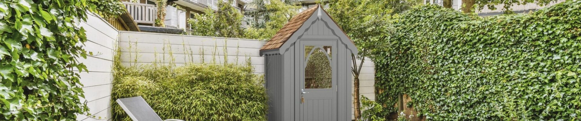 A stylish garden shed, named The Ludlow Shed by The Posh Shed Company, priced at £3999. The shed is grey with a red roof and features a window and a door, situated in a contemporary patio garden.