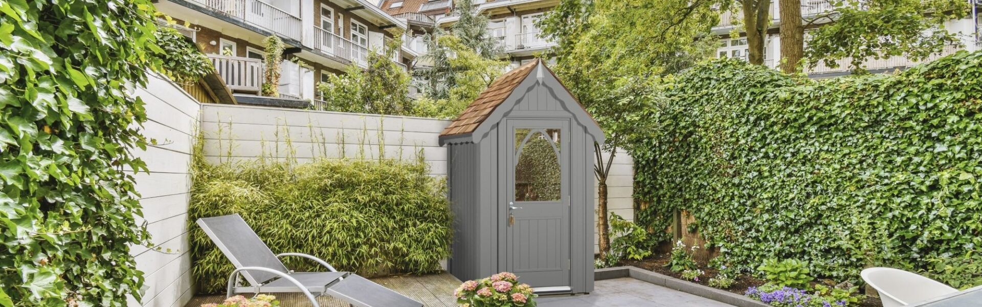 A stylish garden shed, named The Ludlow Shed by The Posh Shed Company, priced at £3999. The shed is grey with a red roof and features a window and a door, situated in a contemporary patio garden.
