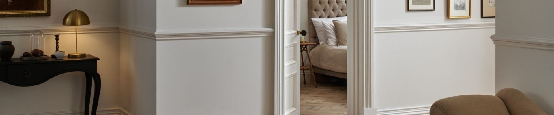 A stylish room featuring Woodpecker Flooring's Orkney Pewter Oak Herringbone Engineered Wood Flooring. The flooring is laid in a classic herringbone pattern, with varying shades of light brown and grey tones. The walls are painted in a light, neutral colour with white wainscoting and moulding.