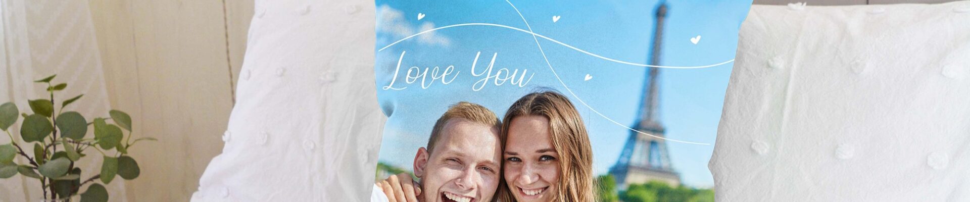 A custom photo cushion on a bed. The cushion features a picture of a smiling couple, with a man on the left embracing a woman on the right. Both are looking directly at the camera. Behind them, the Eiffel Tower is visible against a blue sky with some white decorative swirls and small hearts, and the words "Love You" written in a stylised font.