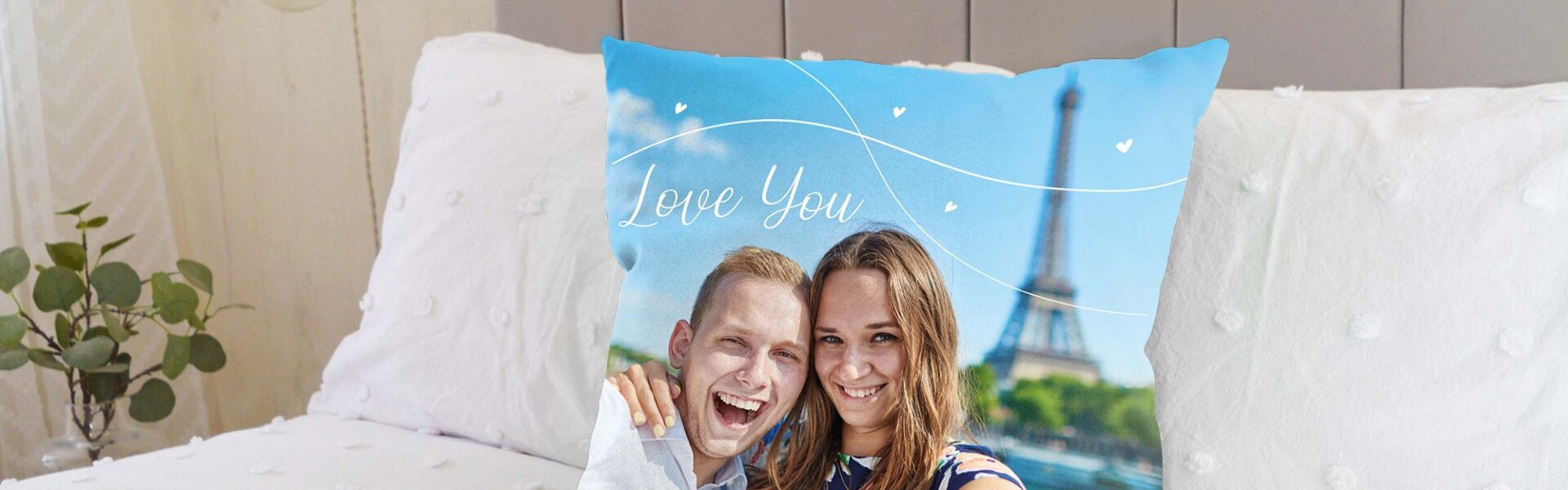 A custom photo cushion on a bed. The cushion features a picture of a smiling couple, with a man on the left embracing a woman on the right. Both are looking directly at the camera. Behind them, the Eiffel Tower is visible against a blue sky with some white decorative swirls and small hearts, and the words "Love You" written in a stylised font.