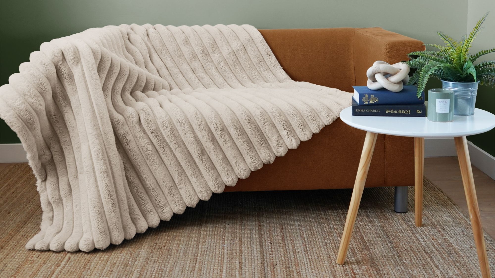 A cosy living room scene featuring a light brown, two-seater sofa. A plush, cream-coloured ribbed faux fur blanket is draped over the sofa, cascading down the front