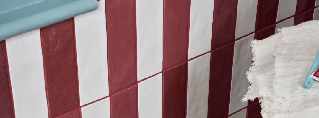 A wall is tiled with alternating vertical stripes of glossy, brick-red and off-white rectangular tiles. The tiles are arranged in a staggered pattern, with each row offset from the one below it. The grout between the tiles is a reddish-brown colour.