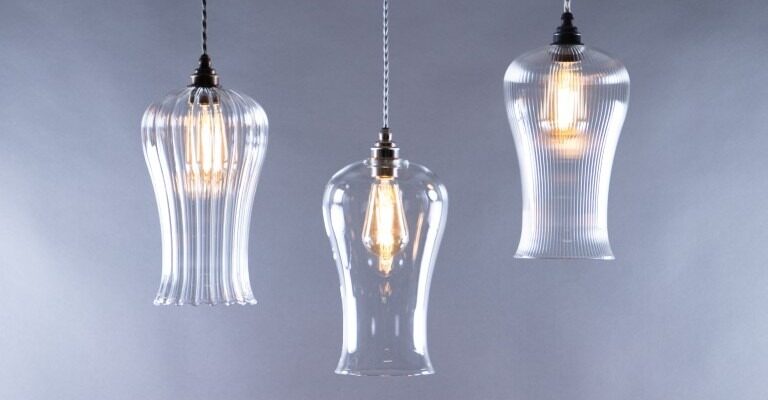 A trio of glass pendant lights hangs against a soft grey background. The lights are suspended by braided fabric cords. The two outer fixtures are identical, featuring a ribbed, fluted glass shade that flares out at the bottom. The central light has a smooth, clear glass shade with a similar bell-like shape.