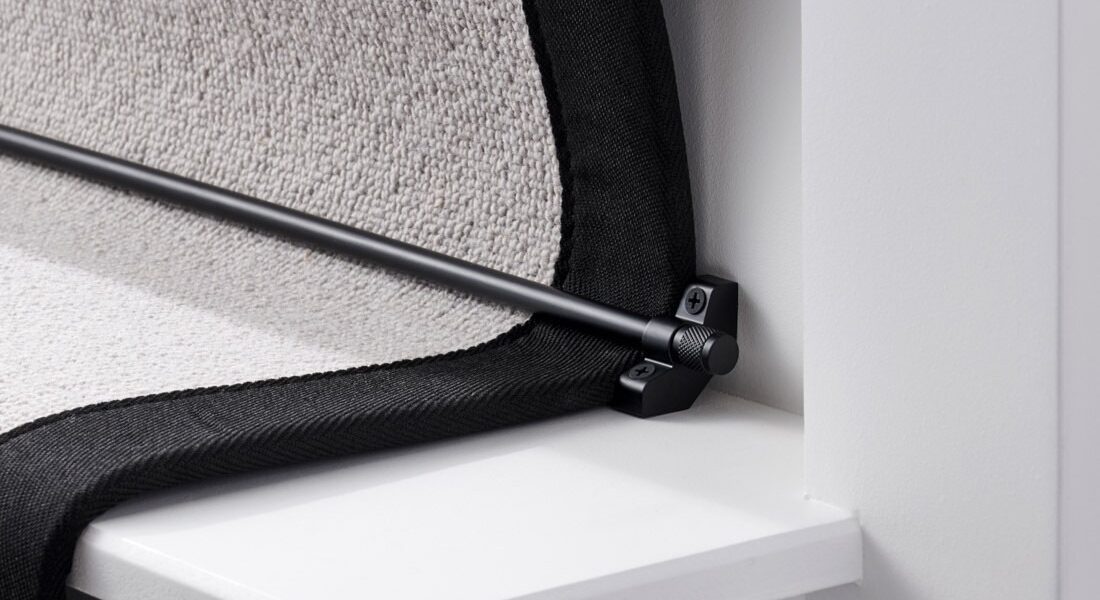 A close-up, low-angle shot of a black stair carpet runner rod installed on a white staircase. The black runner rod is held in place by a matching black bracket on the left, which is screwed into the white stair riser. The bracket has a decorative knob on the front, and the screw is visible.