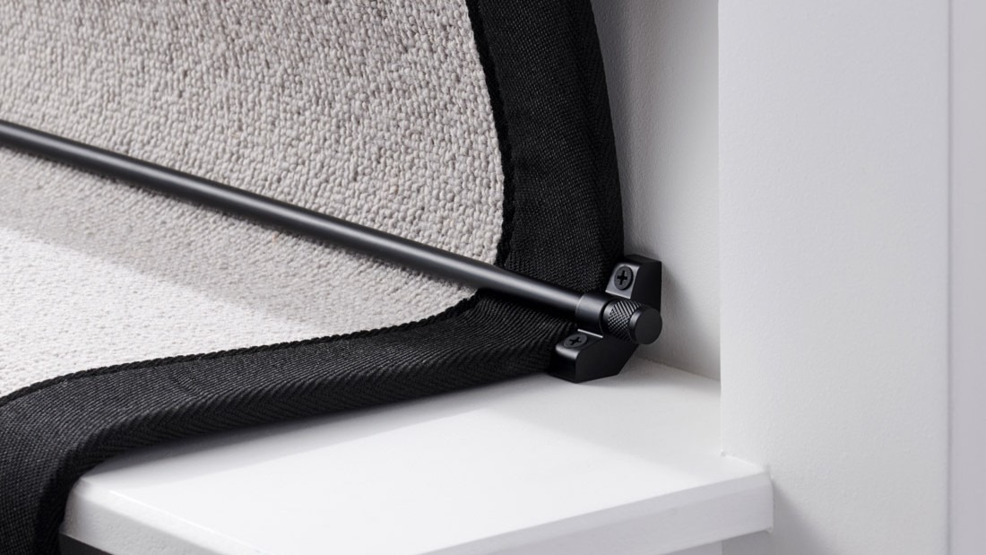 A close-up, low-angle shot of a black stair carpet runner rod installed on a white staircase. The black runner rod is held in place by a matching black bracket on the left, which is screwed into the white stair riser. The bracket has a decorative knob on the front, and the screw is visible.