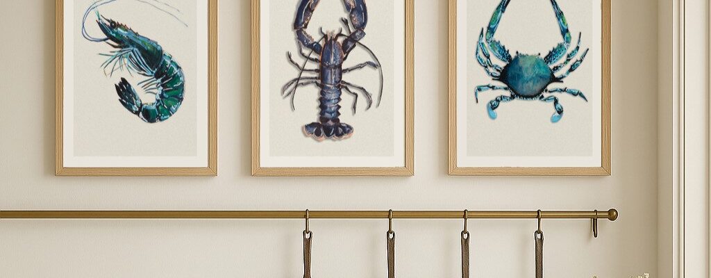 A set of three framed art prints depicting shellfish, including a shrimp, a lobster, and a crab, are displayed in a row on a light-coloured wall above a kitchen counter. Below the prints, there's a brass rail with four copper pans hanging from hooks.