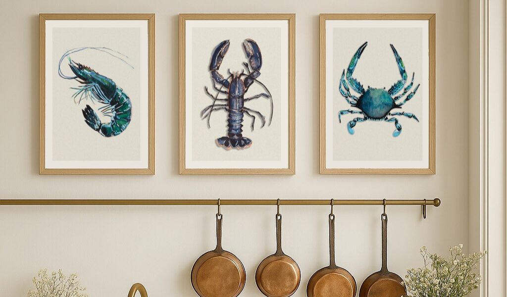 A set of three framed art prints depicting shellfish, including a shrimp, a lobster, and a crab, are displayed in a row on a light-coloured wall above a kitchen counter. Below the prints, there's a brass rail with four copper pans hanging from hooks.