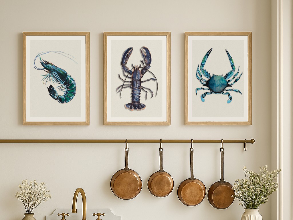 A set of three framed art prints depicting shellfish, including a shrimp, a lobster, and a crab, are displayed in a row on a light-coloured wall above a kitchen counter. Below the prints, there's a brass rail with four copper pans hanging from hooks.