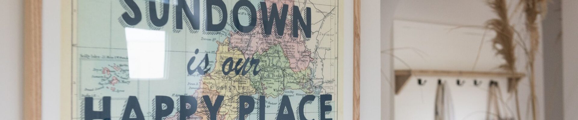 A framed map of Cornwall and the English Channel hangs on a white wall. The map is vintage-style with pastel colours. The text "SUNDOWN is our HAPPY PLACE" is overlaid in bold, black letters on the map