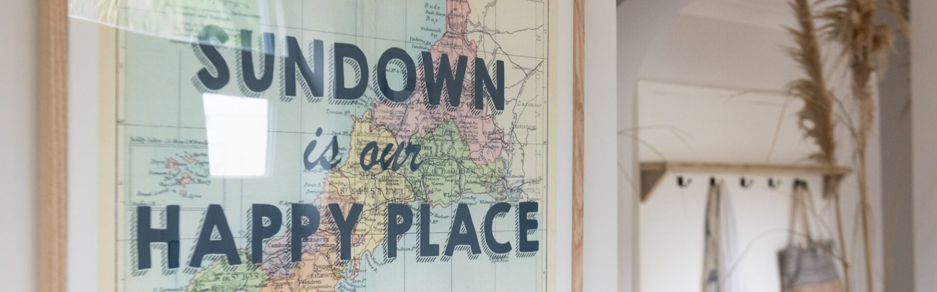 A framed map of Cornwall and the English Channel hangs on a white wall. The map is vintage-style with pastel colours. The text "SUNDOWN is our HAPPY PLACE" is overlaid in bold, black letters on the map