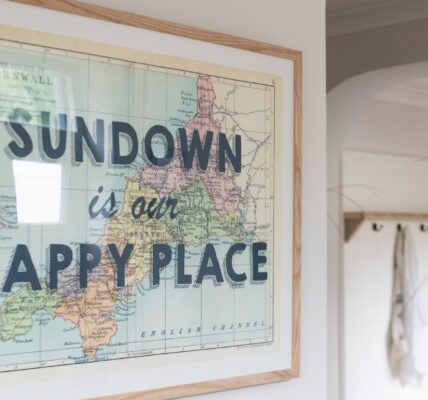 A framed map of Cornwall and the English Channel hangs on a white wall. The map is vintage-style with pastel colours. The text "SUNDOWN is our HAPPY PLACE" is overlaid in bold, black letters on the map