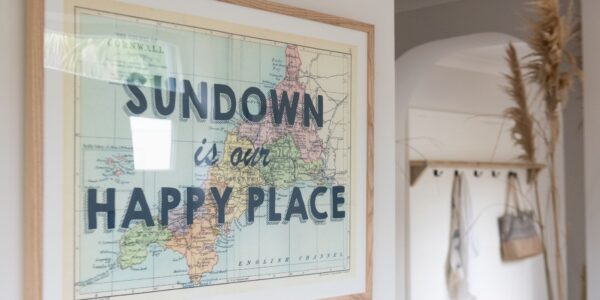 A framed map of Cornwall and the English Channel hangs on a white wall. The map is vintage-style with pastel colours. The text "SUNDOWN is our HAPPY PLACE" is overlaid in bold, black letters on the map
