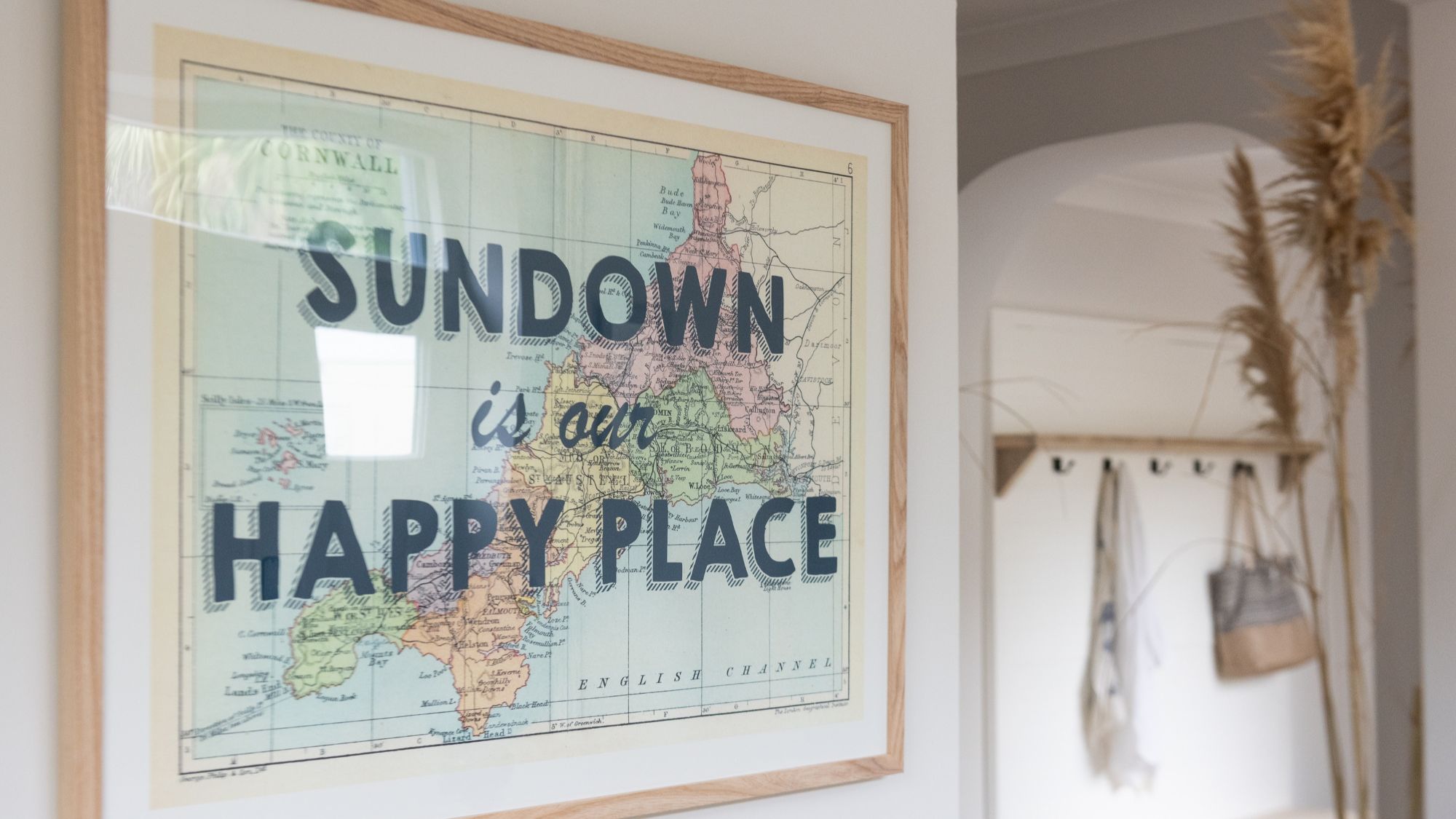 A framed map of Cornwall and the English Channel hangs on a white wall. The map is vintage-style with pastel colours. The text "SUNDOWN is our HAPPY PLACE" is overlaid in bold, black letters on the map