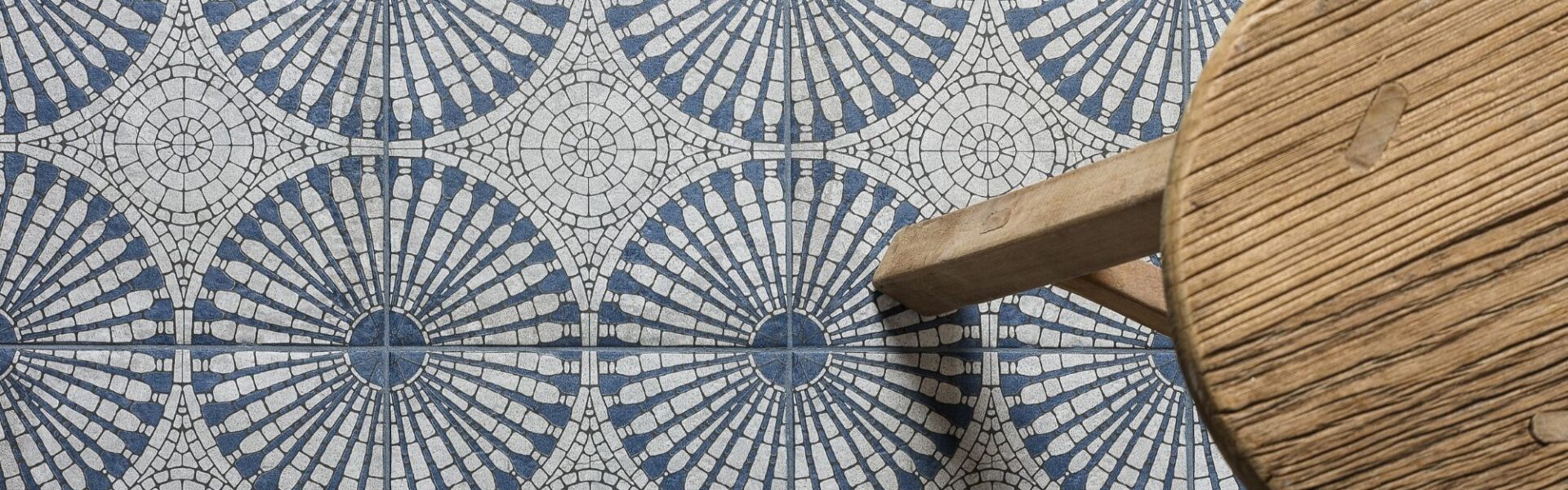 A bird’s eye view of a patterned floor tile. The light grey and blue tile has a circular geometric pattern. A small wooden stool is also seen.