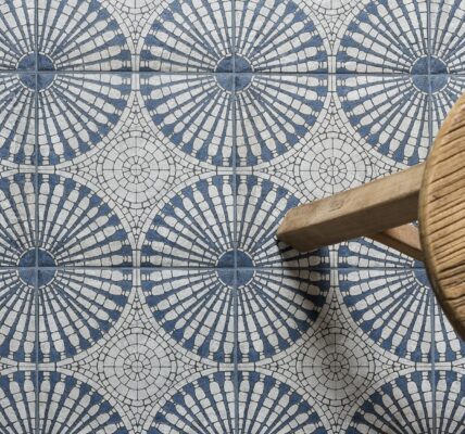 A bird’s eye view of a patterned floor tile. The light grey and blue tile has a circular geometric pattern. A small wooden stool is also seen.