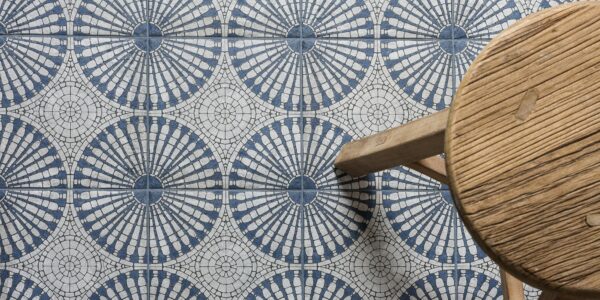 A bird’s eye view of a patterned floor tile. The light grey and blue tile has a circular geometric pattern. A small wooden stool is also seen.