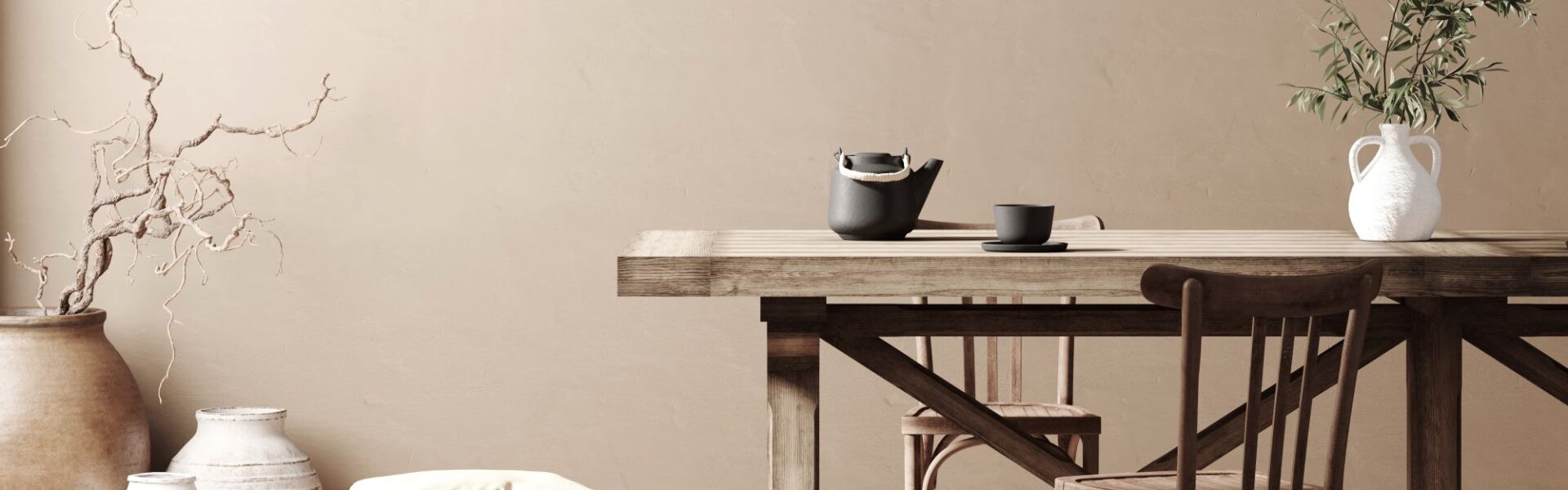 A dining room featuring a long, natural wood dining table and chairs. The walls are a warm, light taupe colour. On the left, a collection of large, textured vases hold decorative branches.