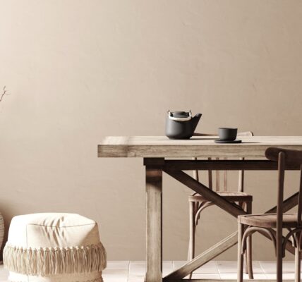 A dining room featuring a long, natural wood dining table and chairs. The walls are a warm, light taupe colour. On the left, a collection of large, textured vases hold decorative branches.