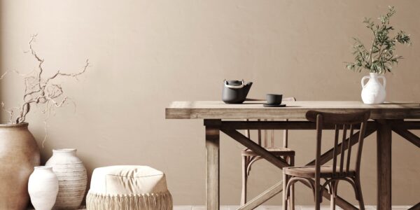 A dining room featuring a long, natural wood dining table and chairs. The walls are a warm, light taupe colour. On the left, a collection of large, textured vases hold decorative branches.