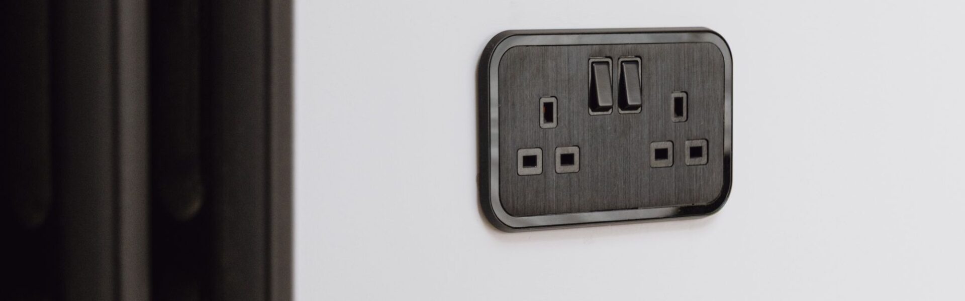 A close-up shot of a UK double-gang plug socket. It's finished in a sleek, dark colour, possibly black or a very dark grey, with a brushed metal texture on the faceplate. The socket is mounted on a plain white wall. There's also a dark radiator visible on the left side of the image.
