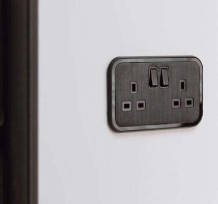 A close-up shot of a UK double-gang plug socket. It's finished in a sleek, dark colour, possibly black or a very dark grey, with a brushed metal texture on the faceplate. The socket is mounted on a plain white wall. There's also a dark radiator visible on the left side of the image.