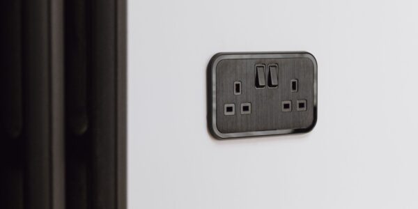 A close-up shot of a UK double-gang plug socket. It's finished in a sleek, dark colour, possibly black or a very dark grey, with a brushed metal texture on the faceplate. The socket is mounted on a plain white wall. There's also a dark radiator visible on the left side of the image.