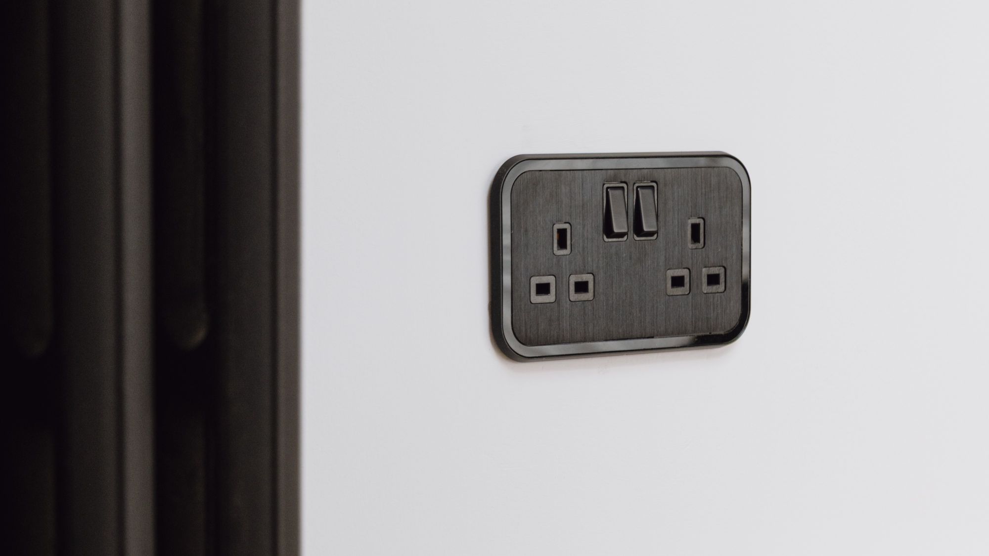 A close-up shot of a UK double-gang plug socket. It's finished in a sleek, dark colour, possibly black or a very dark grey, with a brushed metal texture on the faceplate. The socket is mounted on a plain white wall. There's also a dark radiator visible on the left side of the image.