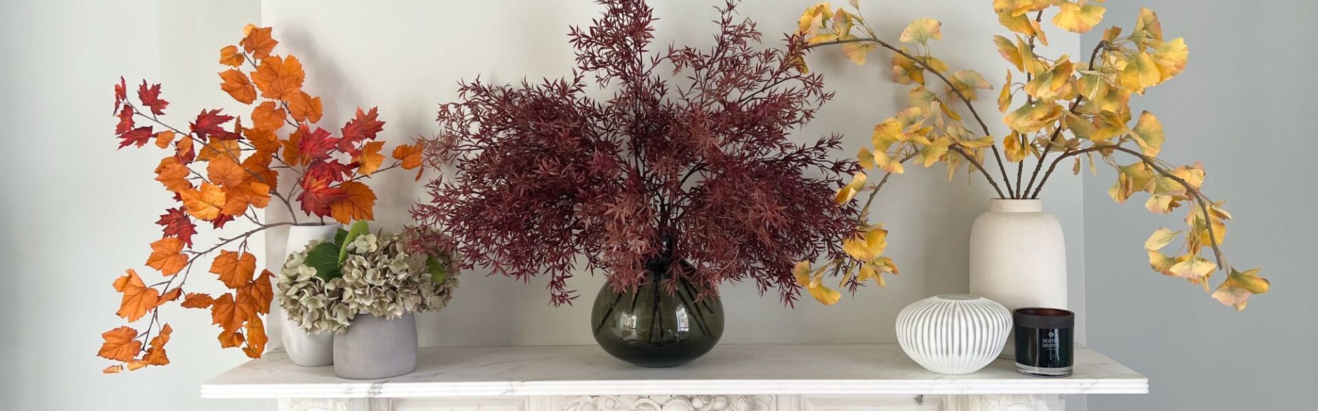 A white marble fireplace is decorated with three vases of artificial autumnal leaves. A small bunch of realistic-looking grey-green hydrangeas sits next to the vase on the far left. The vase on the left holds bright orange maple leaves on a branch. The centre vase holds a sprawling, deep burgundy-coloured branch with fine, fern-like leaves.