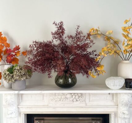 A white marble fireplace is decorated with three vases of artificial autumnal leaves. A small bunch of realistic-looking grey-green hydrangeas sits next to the vase on the far left. The vase on the left holds bright orange maple leaves on a branch. The centre vase holds a sprawling, deep burgundy-coloured branch with fine, fern-like leaves.